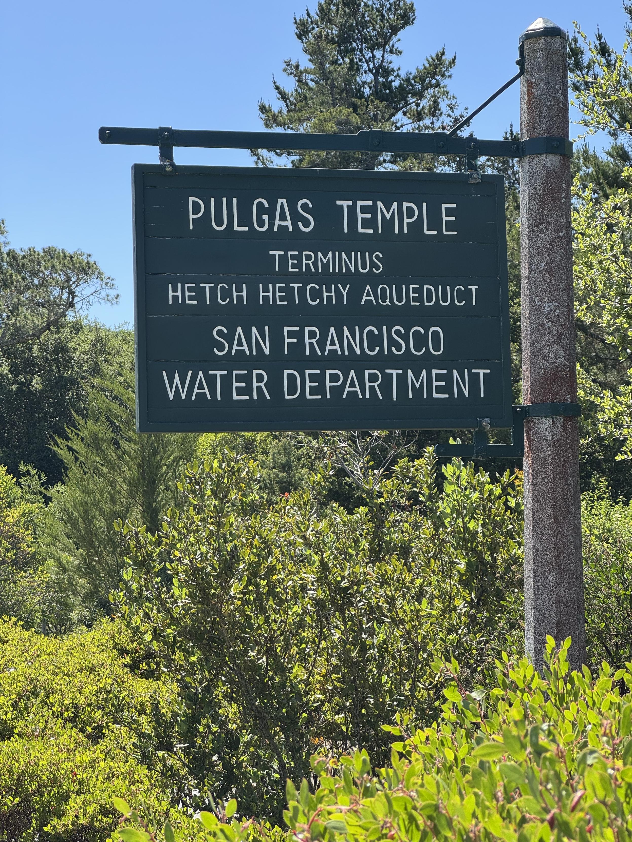 A green sign with white uppercase routed letters hanging from a green pipe on a larger steel pole, surrounded by green deciduous, pine, and bushes under a clear blue sky, which reads:PULGAS TEMPLETERMINUSHETCH HETCHY AQUEDUCTSAN FRANCISCOWATER DEPARTMENT