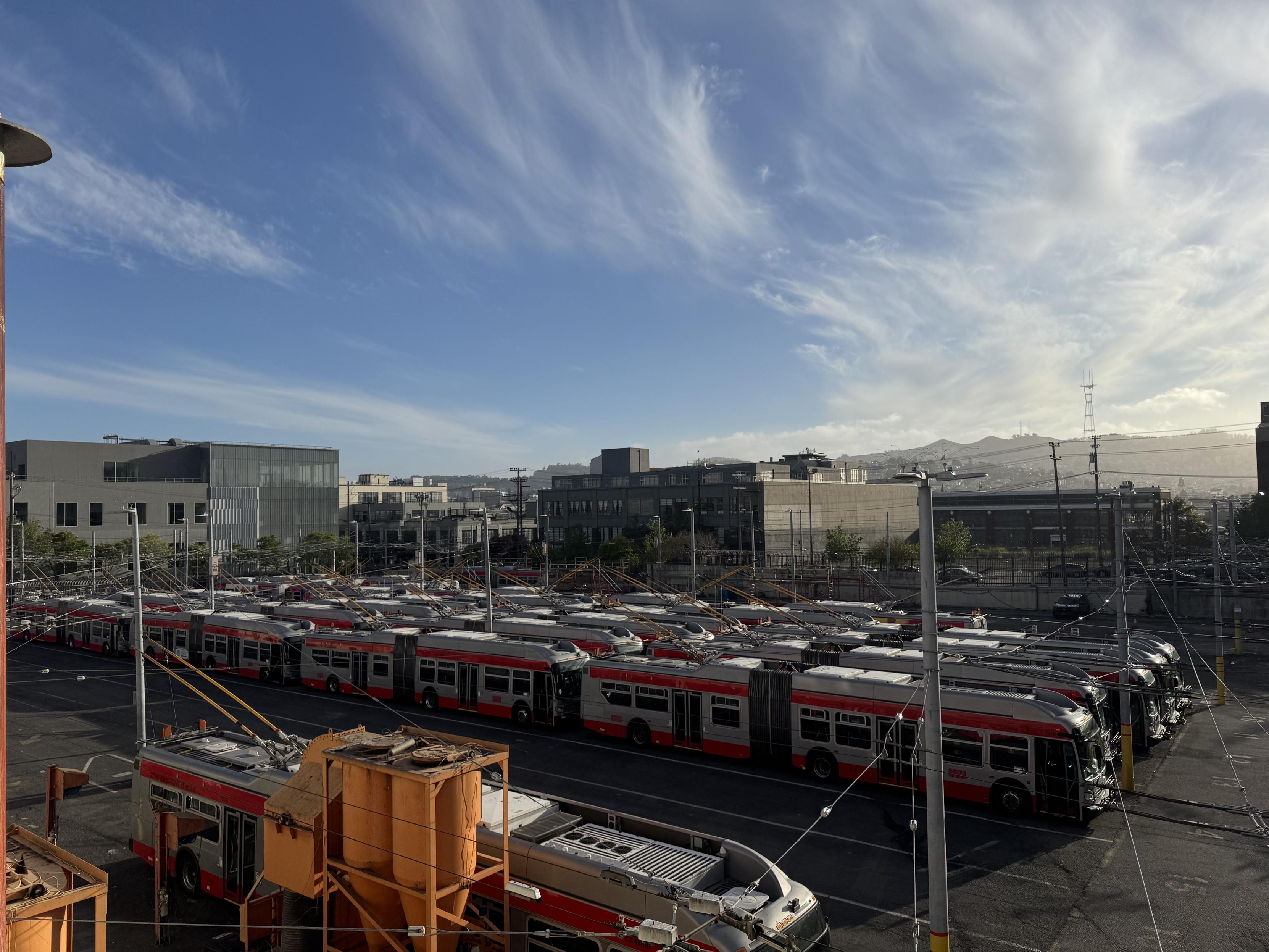 A photograph looking southwest from the rooftop of the yard where the bus tire shed is, looking down at buses that remain at the full capacity station when most are out on routes. Dozens of SF MTA trolley busses are parked in rows in the foreground, with the closest parked next to what I learned are giant industrial vacuums that are not used anymore. The top half of the photo is a blue sky with whispy white clouds. Sutro tower, an SF landmark, is recognizable on the horizon to the right.