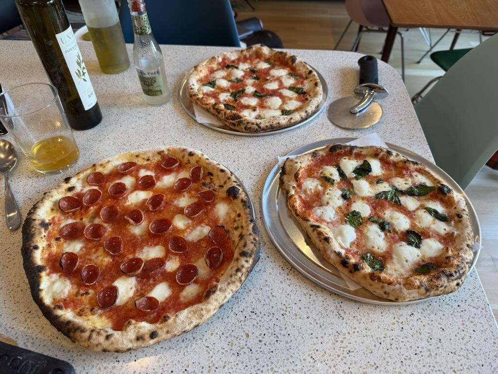 three uncut 12” neapolitan pizzas: two margheritas and one pepperoni, on a counter with a pizza peel, two kinds of olive oil, a glass of beer, and a bottle of ginger ale
