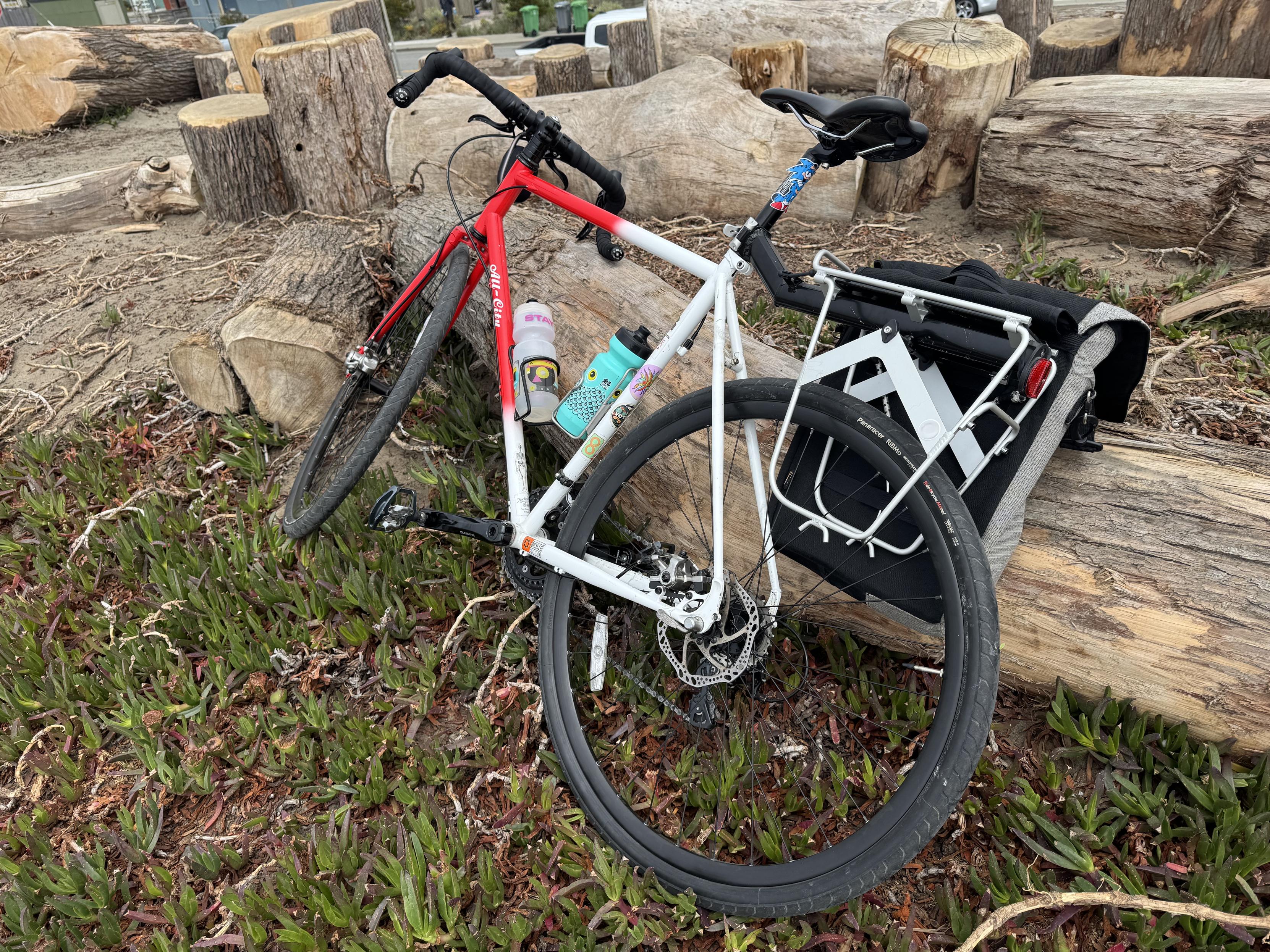 my bike, rolled up off the path onto the ice plant, leaning against a logit’s an ~10 year old gravel bike w/ slicks on it (an all-city macho man with disc breaks and 105 components) red on the front half and white on the back, with a buncha stupid stickers on the white half. I’ve got two water cages on it use both, a panier rack and bag.