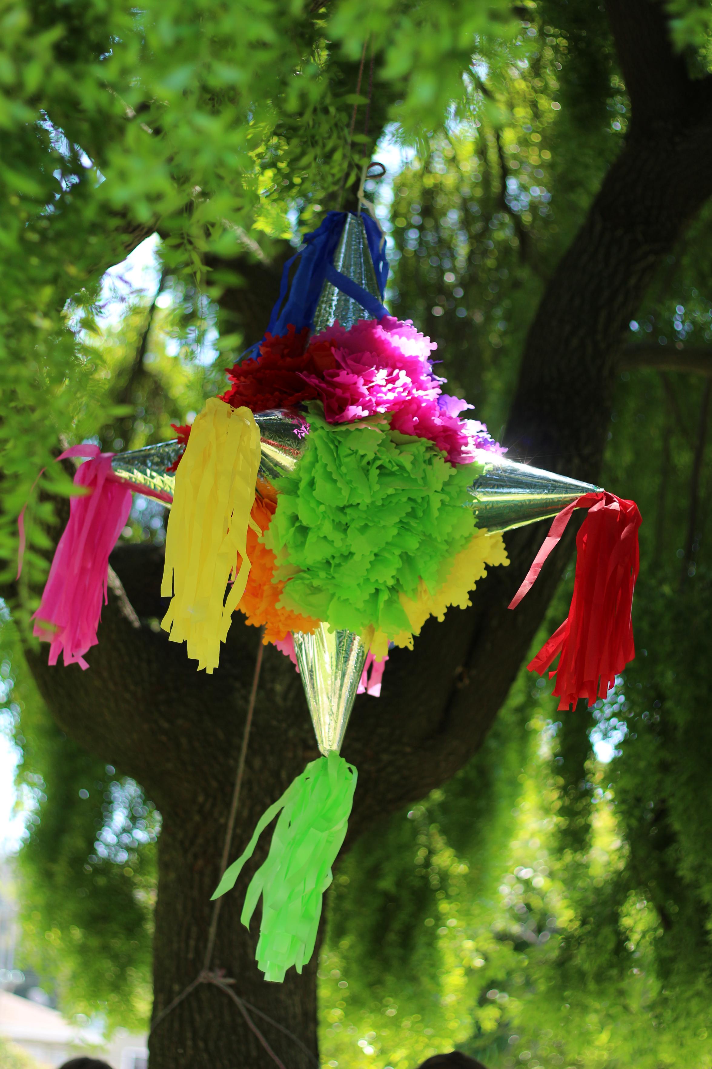 a rainbow ball piñata with silver reflective party hates taped all around it with ribbon tassels on the ends hanging from a tree