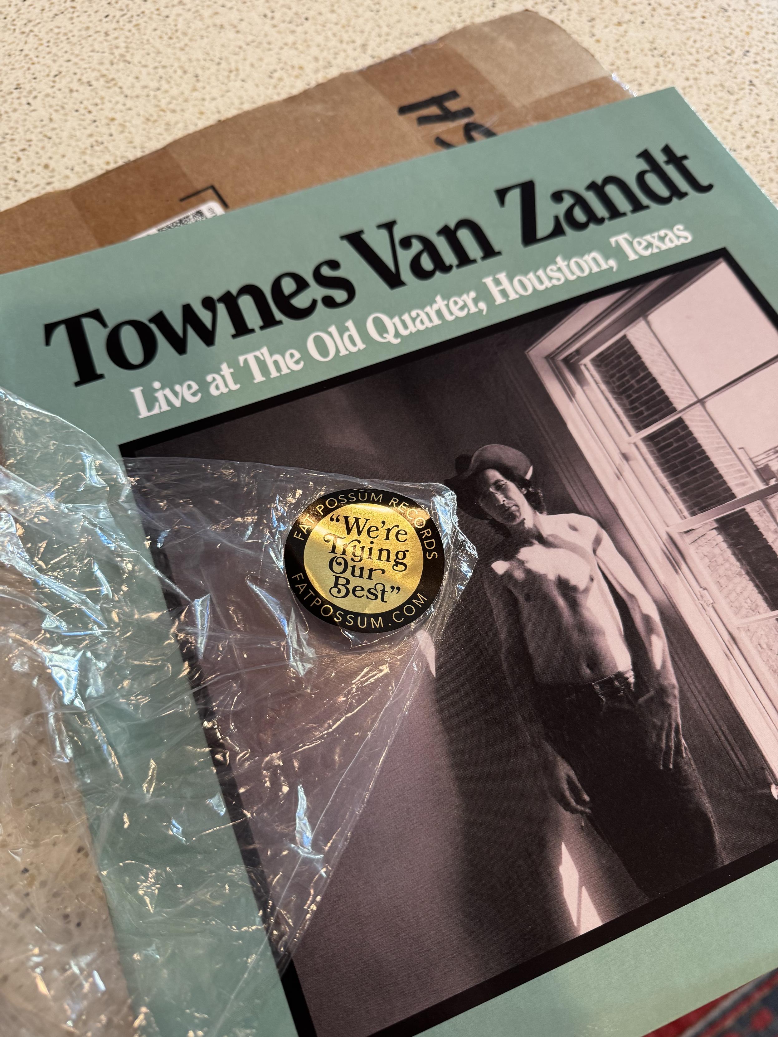 Pic of a record I just took out of the carboard shipping box it’s laying on, on my kitchen island. Townes Van ZandtLive at The Old Quarter, Houston, TexasI ripped the clear plastic shrink wrap off as per, but laid it back on to get a pic of the label’s round sticker on it, which proclaims FAT POSSUM RECORDS and their .com in gold on black around the outter edge, and “We’re Trying Our Best” in a cute retro black on gold in the center, with the grocer’s quotes and everything.