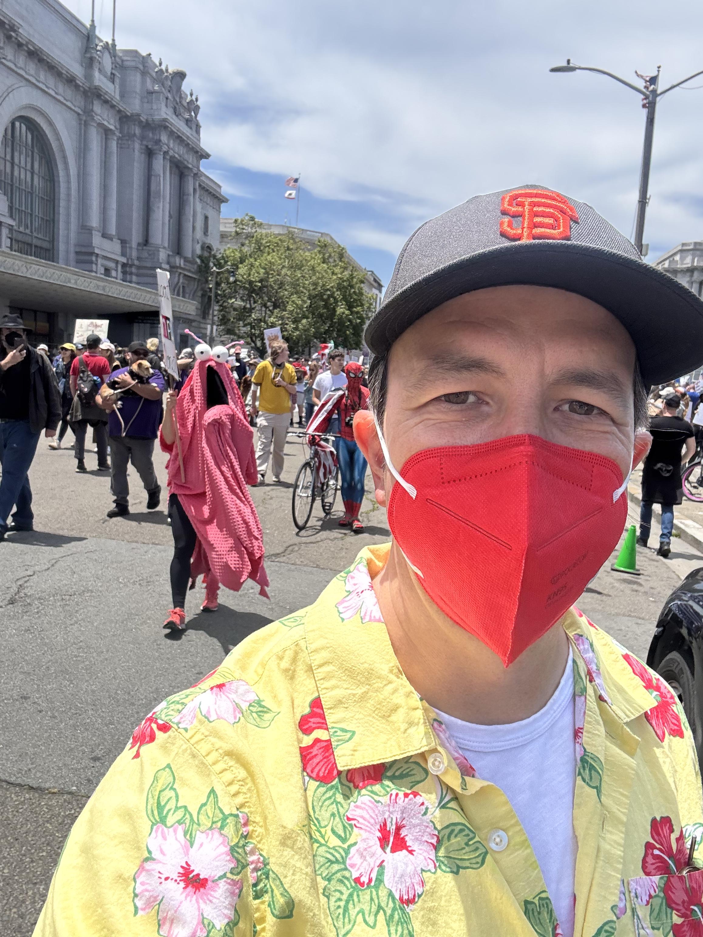 selfie, man (me) in a yellow camp shirt w/ flowers on it, red kn95 mask and black SF giants hat. behind me someone with a “save NPR” sign is dressed like the martians from sesame street who panic every time the phone rings trying to figure out what it isfurther in the distance someone in a spiderman costume pushes a bike