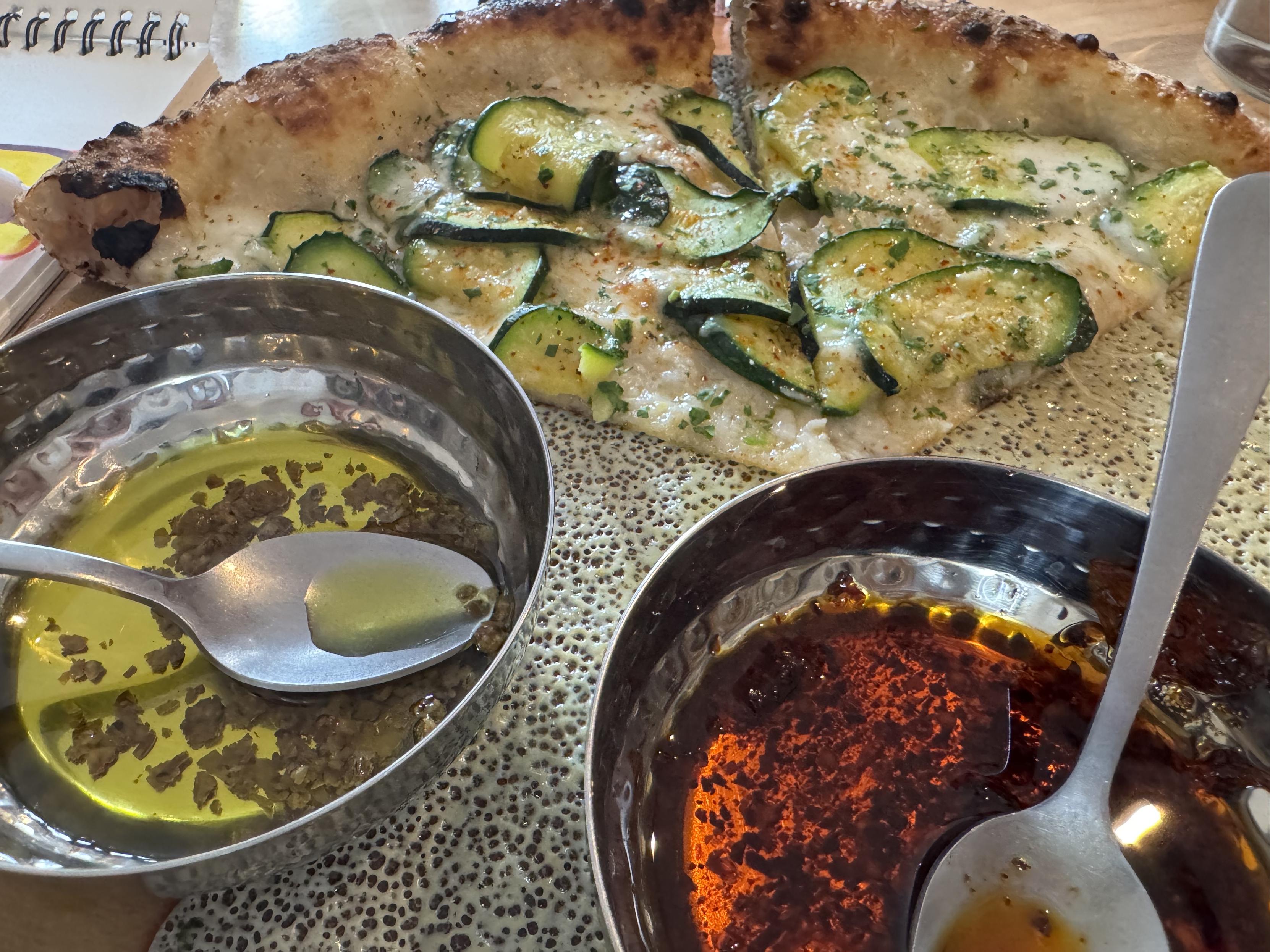 two slices of neapolitan pizza topped with mozzerella and thin slices of zucchini, the crust is salted with maldon or similar large flakes of salt, and leopard spotted perfectlyin the foreground are two stainless steel hammered dishes of chili oil, one green, one red