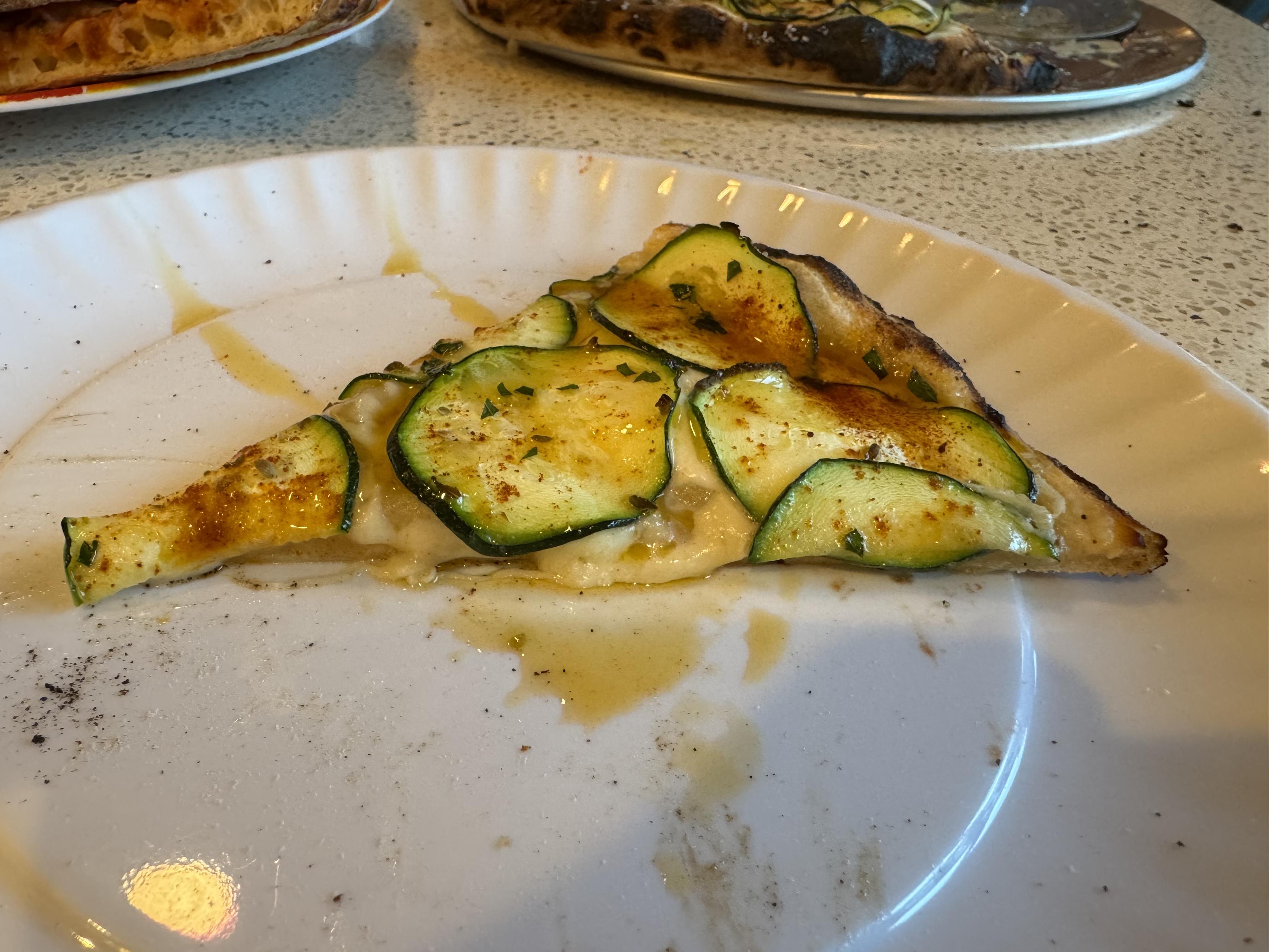 a slice of neapolitan pizza on a fake paper plate made of melamine, drizzled with chili oilthe pizza is topped with micro-planed parmesan, smoked mozz, low moisture mozz, and fresh mozz over which are layered the thinly slice zucchini, smoked paprika, and minced flat leaf parsley. I salted the corniccione with fleur de sel