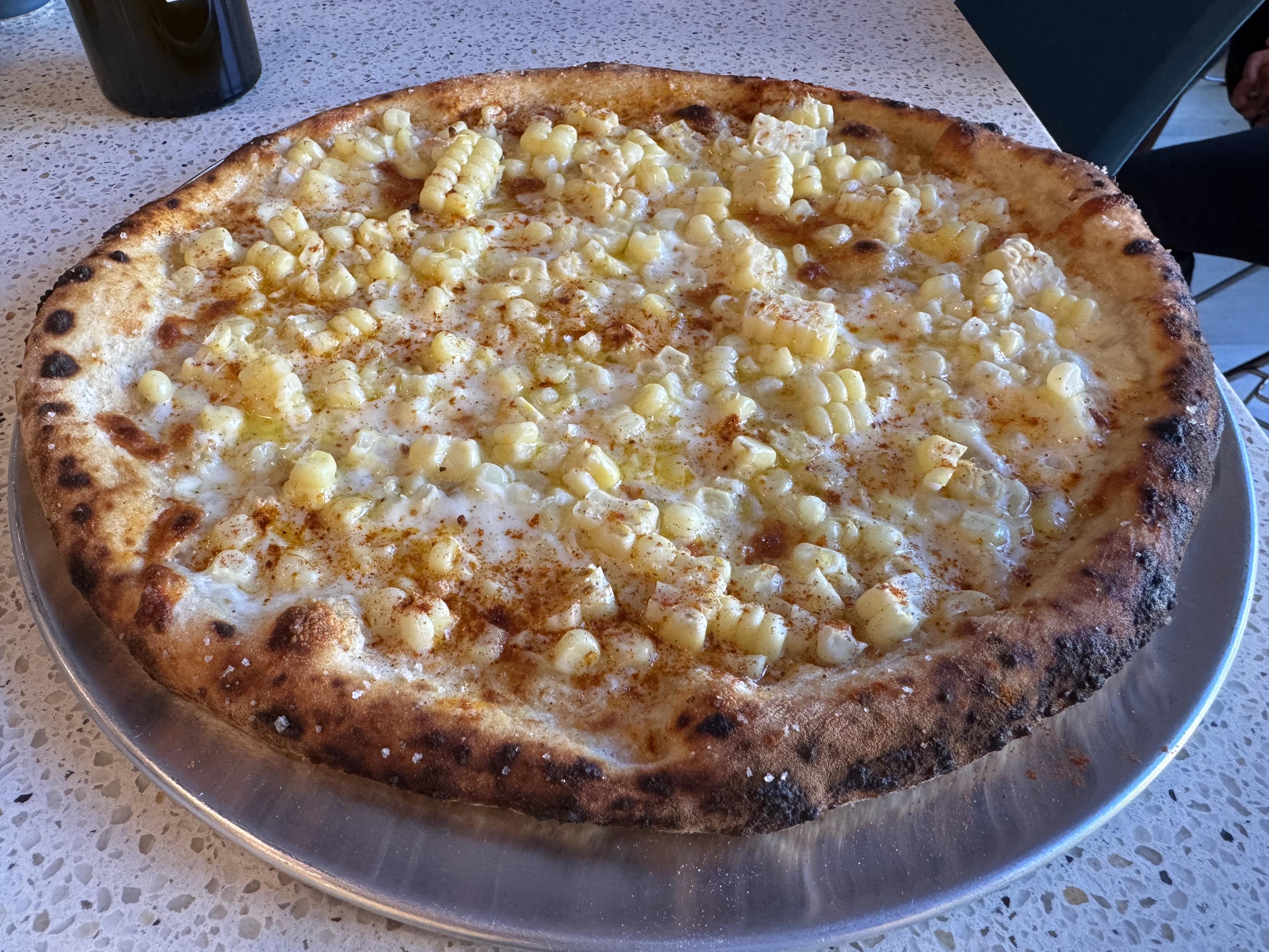 an uncut whole neapolitan pizza on a stainless steel platethe pizza is topped with micro-planed parmesan, smoked mozz, low moisture mozz, and fresh mozz, sweet corn sliced off the cob, and smoked paprika. I salted the corniccione with fleur de sel
