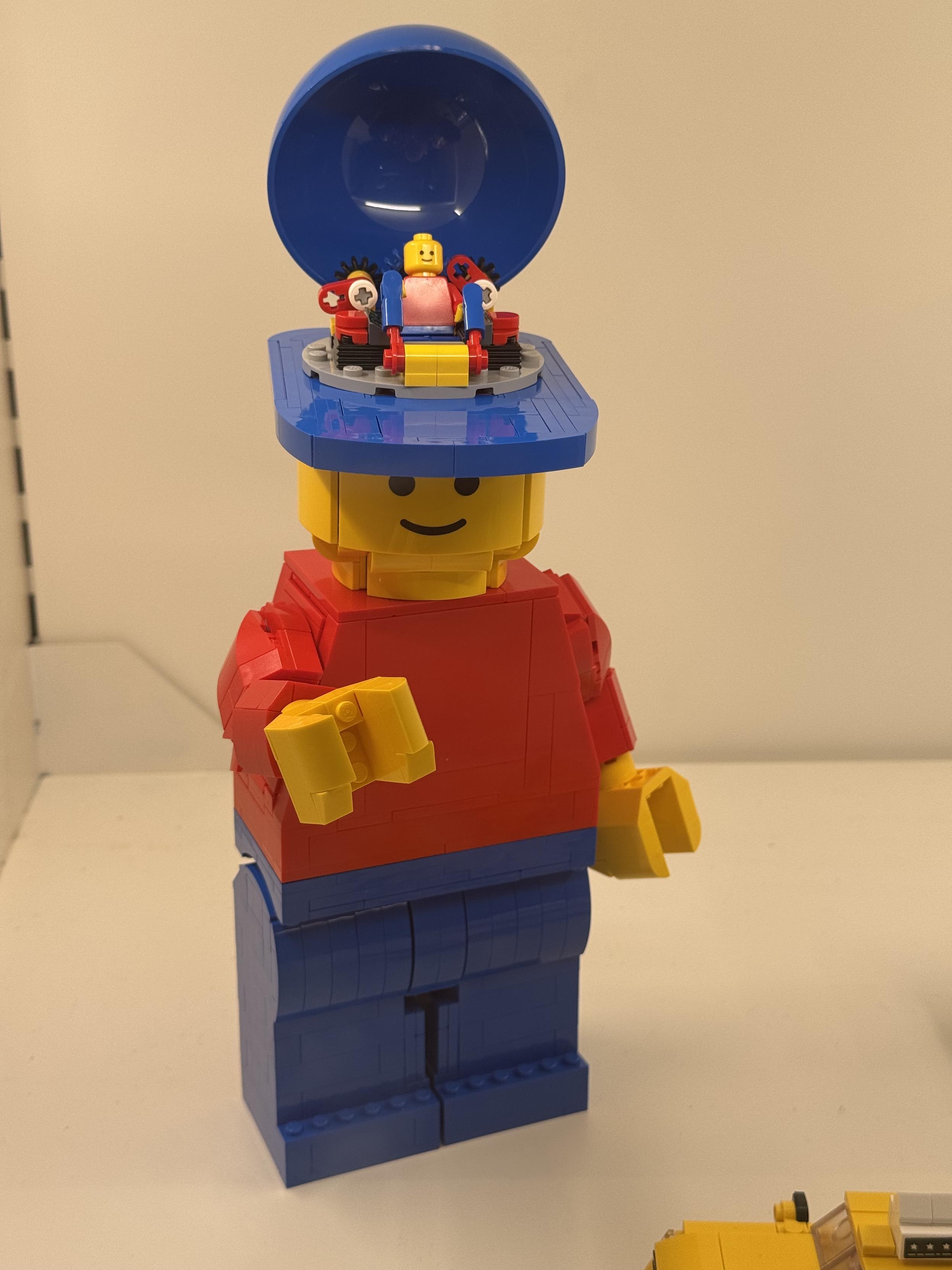 an up-scaled lego minifig, itself made of legos, about 50 bricks tall, wearing a hat which is tilted back to reveal an identical normal sized minifig who is operating the larger version of itself using levers and gears