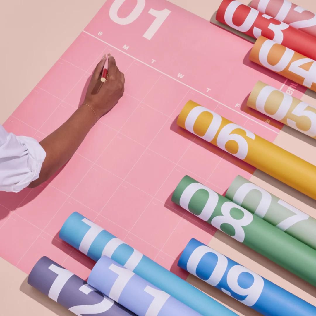an image from poketo’s website advertising poster size monthly calendars which arrive in a roll indicating the month and days of the week but not number of days in white helvetica (or something like it) on solid, pale, color paper, which the buyer can then fill in themselves. Each month is a different color of the rainbow as pictured. I’ve also used similar designs from them with abstract patterns. A hand is writing in marker on something on the first monday of January, which is on pink paper. The rest of the months are visible rolled up to the right with the large month number showing on paper in salmon, red, orange, tan, yellow, pale green, green, blue, light blue, violet, and purple.