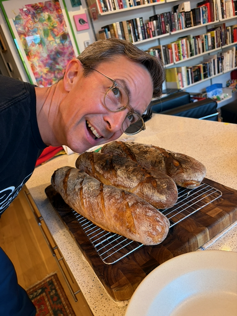 A man (the poster, me) in glasses leaning over, head cocked to one side, putting his ear to three baguettes on a cooling rack on a cutting board. The loaves are fresh from the oven and making ticking, hissing, and cracking noises as they cool.