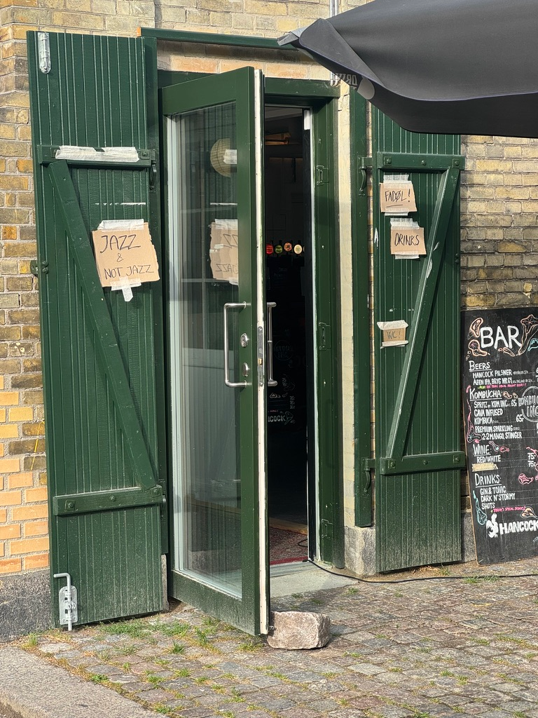 The doorway entrance to a brick building: a steel door fully glazed with a single piece of glass, frame painted green, propped open using a cobblestone, surrounded by wide open wooden shutter doors painted the same green. To the right of the shutters is a chalkboard listing bar specials.A piece of cardboard is taped to the left shutter with masking tape that says “Jazz & Not Jazz.” Smaller pieces taped to the right shutter do that thing bars love to do which, is point out that you can get drinks there (both in Danish and English, “FADØL!”, “DRINKS!”)JAZZNOT JAZZSSADFADØL!ORINKSBARBEERSHANCOCK PILSMER osCAMA FUSEDKOMBUCANAO PREMIUM SERLING E222H4M60, STERWINE:REDIAHITEDRINKS:GIN& TONGDARK M'STURNILSHOTSCHANCOCA