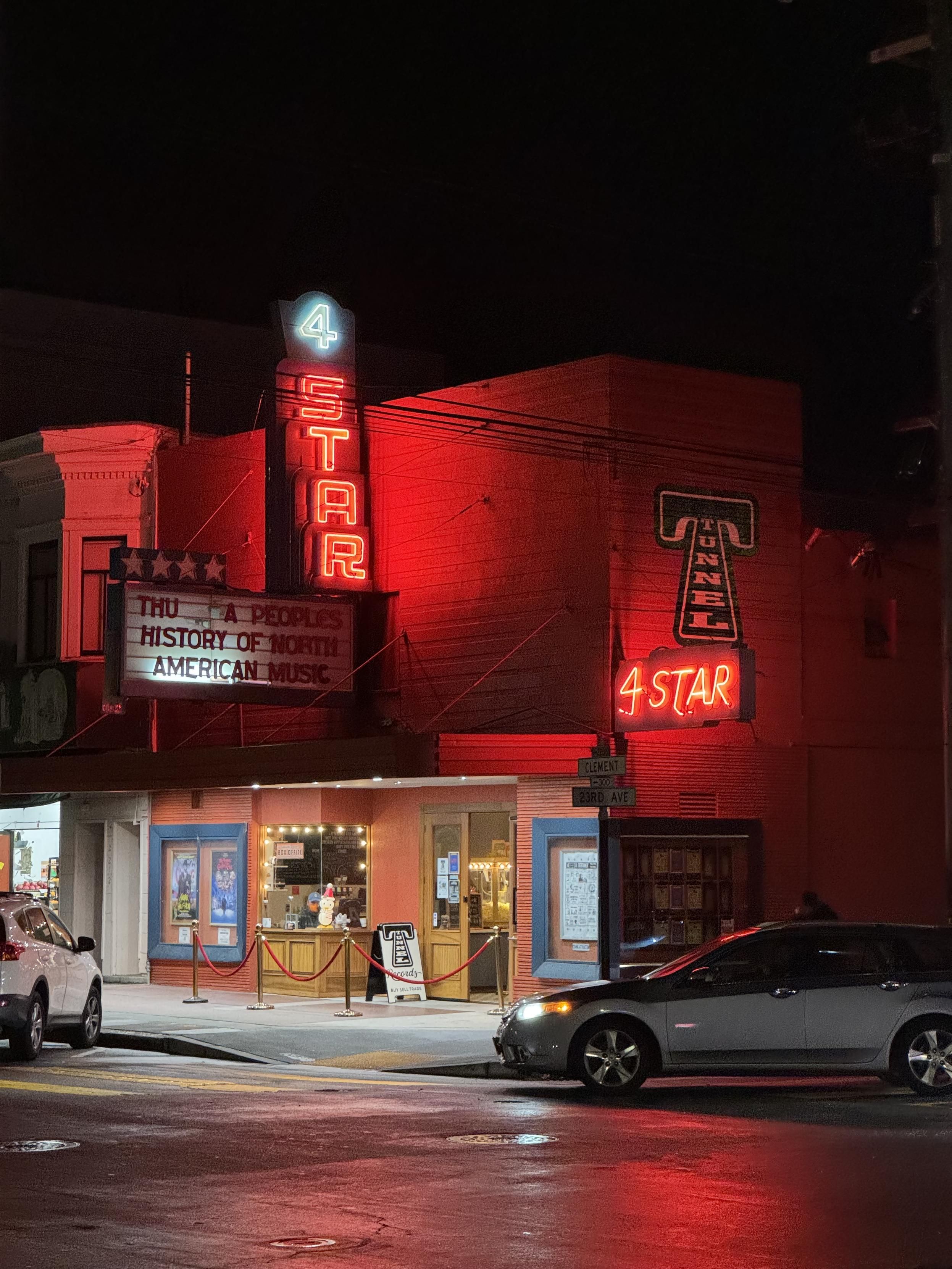 Photograph of the 4 Star theater from kitty-corner on 23rd and Clement, at night. The venue is a 2 or 3 story building with a vertically oriented neon sight of a white star over red letters S, T, A, and R rendered as neon block oblique outlines in front on Clement, and a smaller, lower neon sight on the 23rd that simply says 4STAR in oblique neon linesas a result the top half of the building is washed in red neon glowthe marque is a simple protruding and hanging letterboard with 4 white stars above it, proclaiming tonight: THU: A PEOPLESHISTORY OF NORTHAMERICAN MUSICThe entrance winds past a box office window to a door near the corner brightly lit from above