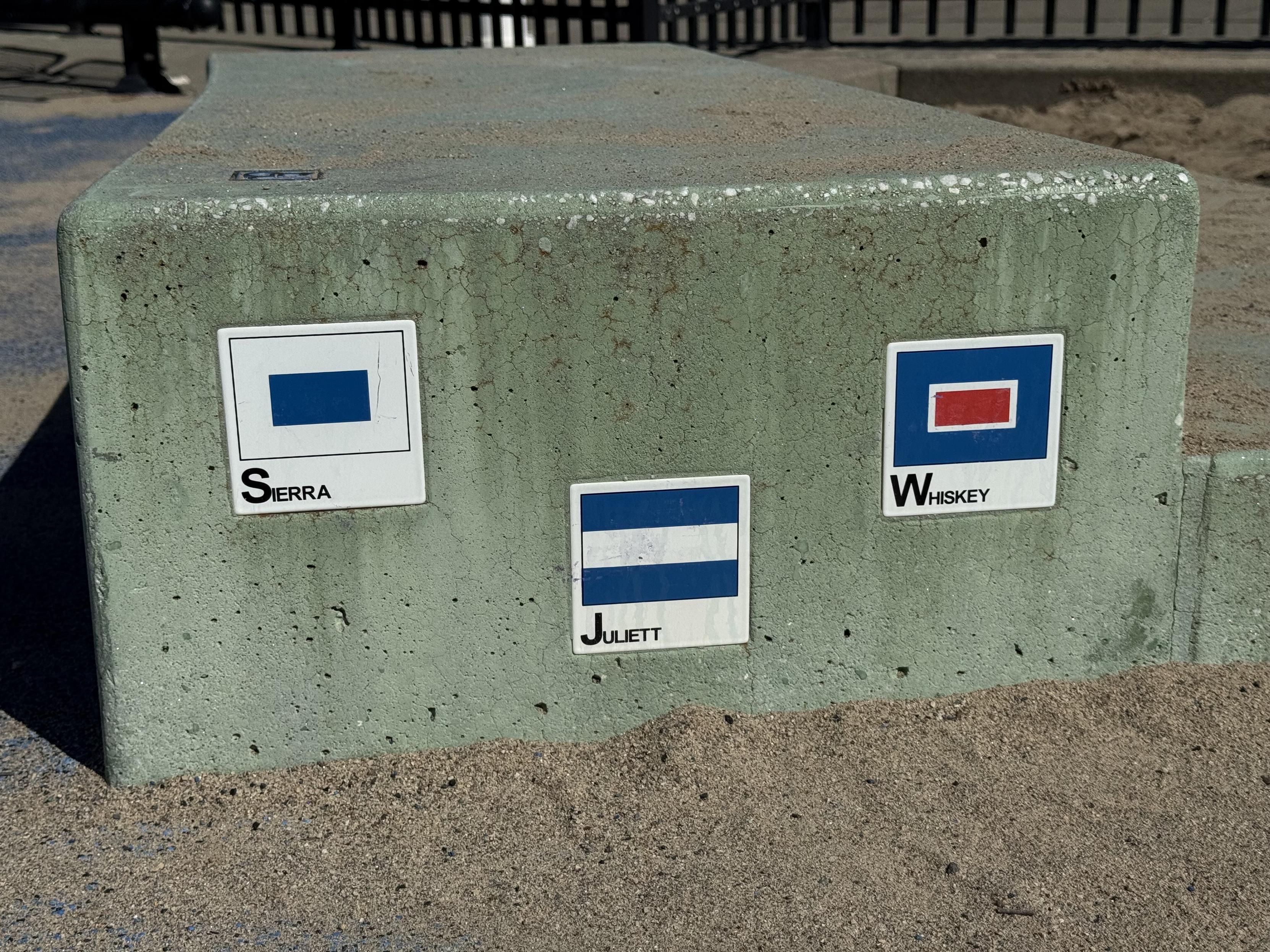 photograph of small nautical flag (i assume?) tiles embedded in concrete steps at a toddler playground in san francisco with Sierra, Juliett, and Whiskey written under each flag, The initial letters are large while the remainder are small caps.