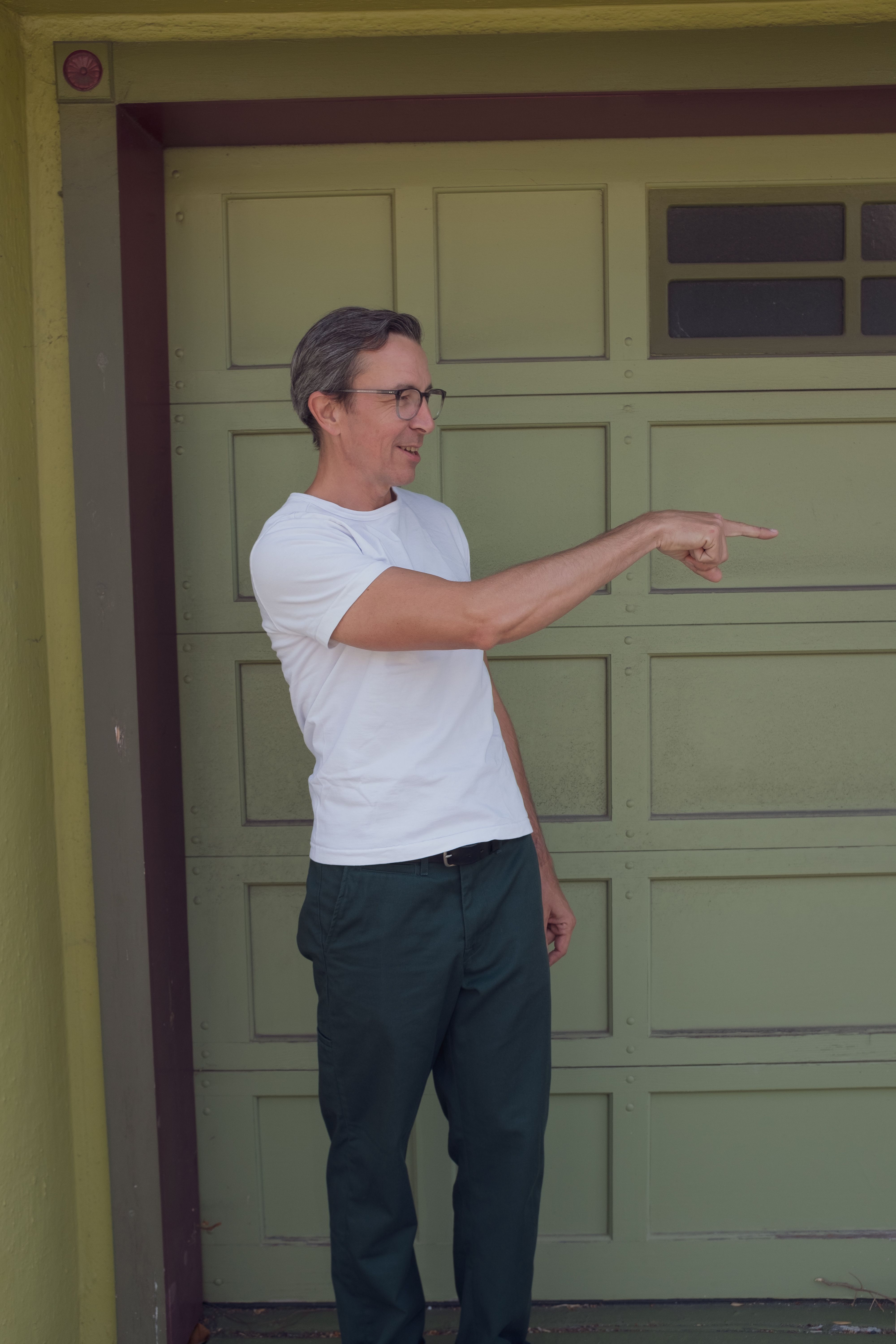 A 35mm-ish potrait of a man (gravely) standing facing the camera wearing dark green work pants and a white t-shirt looking right and pointing right with his right hand. The man is wearing black eyeglasses that fade to clear on the bottom rim. He has a slightly receding hairline and salt and pepper dark hair. We won a school fundraiser raffle with a cool local photographer and this was one of the pictures that came out of a larger set of casual street walk-arounds with the family