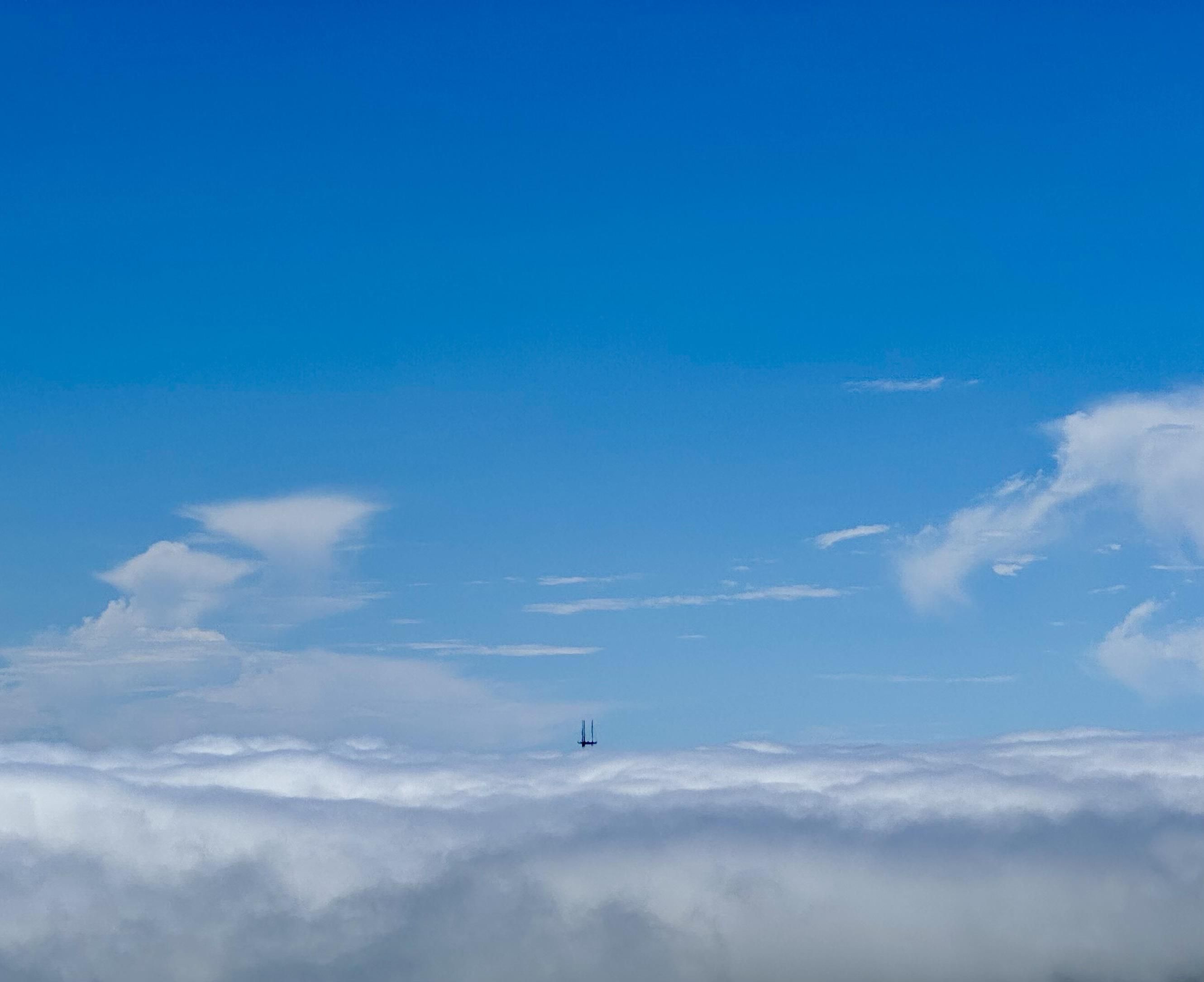 Sutro Tower from Mount Caroline Livermore on Angel Island. The bottom 1/5th of the photo is is a solid layer of clouds. In the center, the tiny “pirate ship” spires of Sutro Tower and the barest bit of the three horizontal braces peeks above the clouds. The rest of the photo is blue sky with a few much higher faint wisps of clouds.