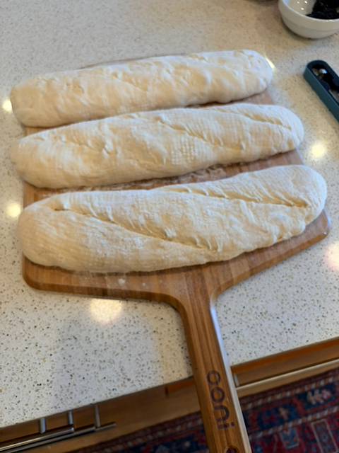 Three loaves of shaped baguette dough, about fifteen inches by four inches, slid on rice flour onto a wooden pizza peel and just before being launched into the oven. Each has been slashed with a razor at a sharp angle, the farthest five times, the middle three times, and the nearest twice.