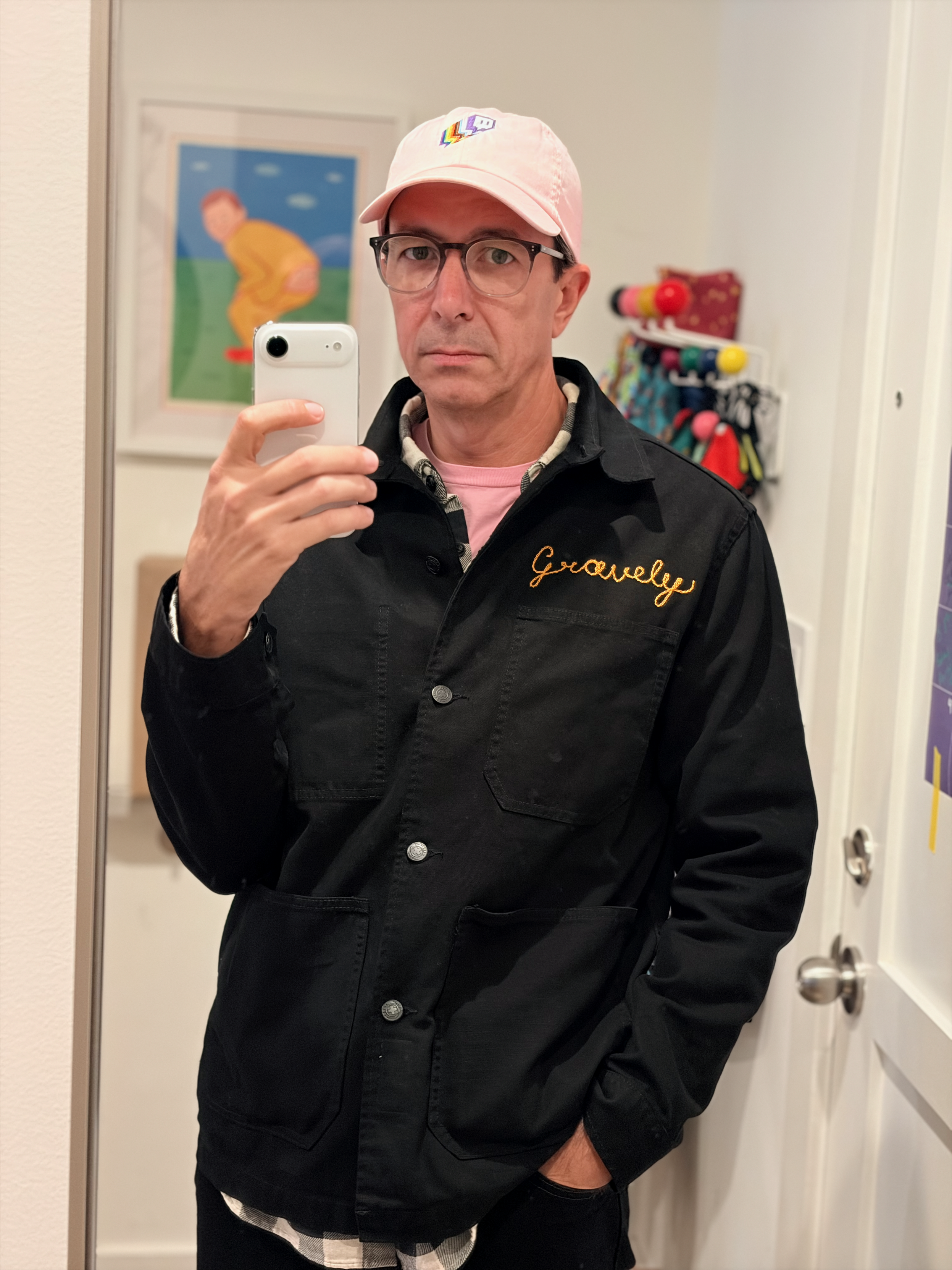 Selfie at home in the mirror by the door. I’m wearing a black 501s and a black chore coat with my handle, Gravely, chain stitched on the right breast in goldenrod thread, over a black and white plaid flannel over a light pink t-shirt. I’m wearing a new-to-me Twitch Unity Guild baseball hat in light pink with the twitch glitch logo with stacked unity pride flag drop shadows. A blurred print of Booty-Boop, my old avatar, by Joan Cornella is behind me on the wall over one shoulder and a rainbow Eames ball coat rack on the wall over the other shoulder.