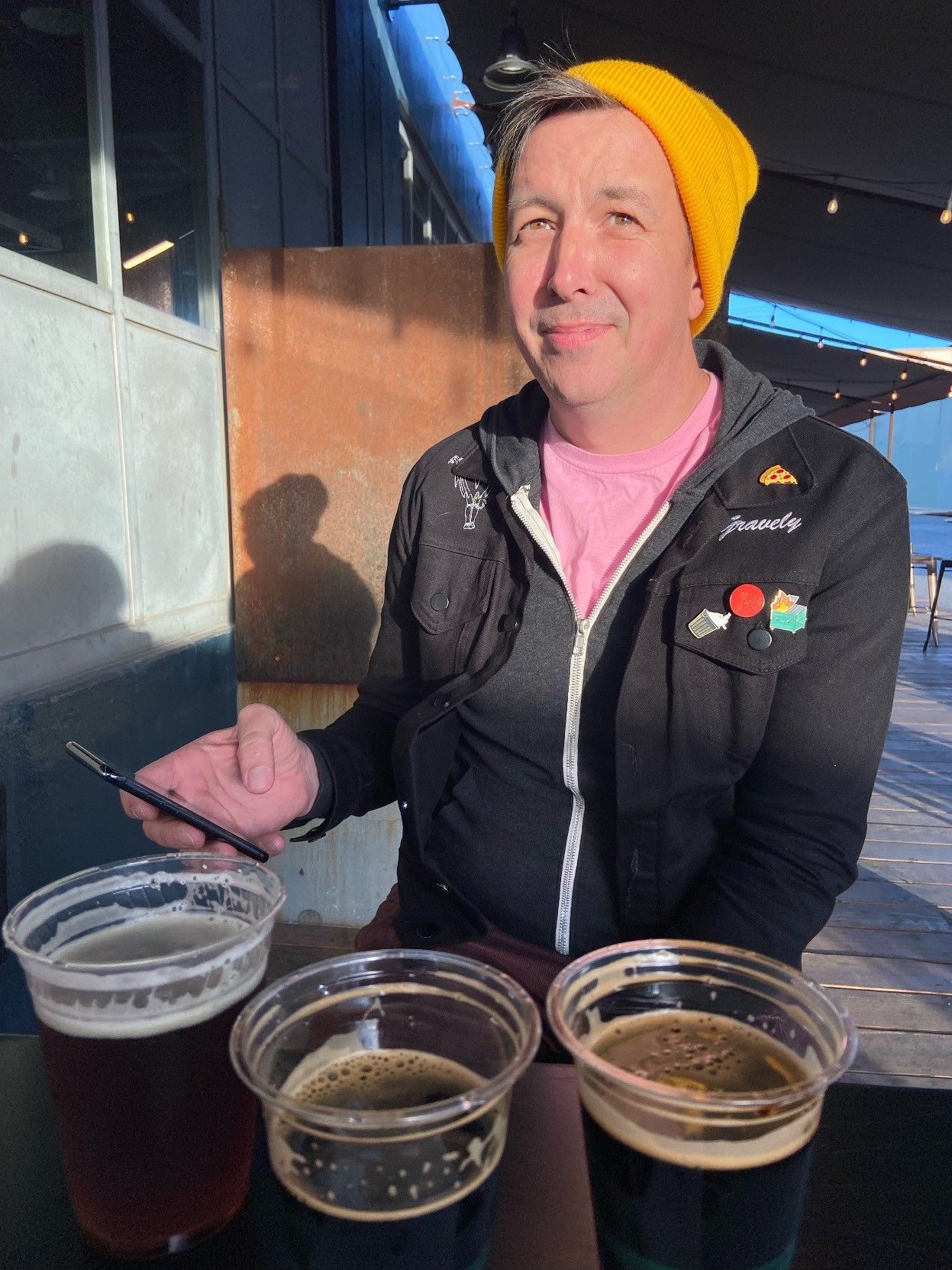 a picture of me squinting into the sun wearing a yellow hat, pink shirt, grey hoodie, and black jean jacket, phone in hand, with a round of beers on the table in front of me