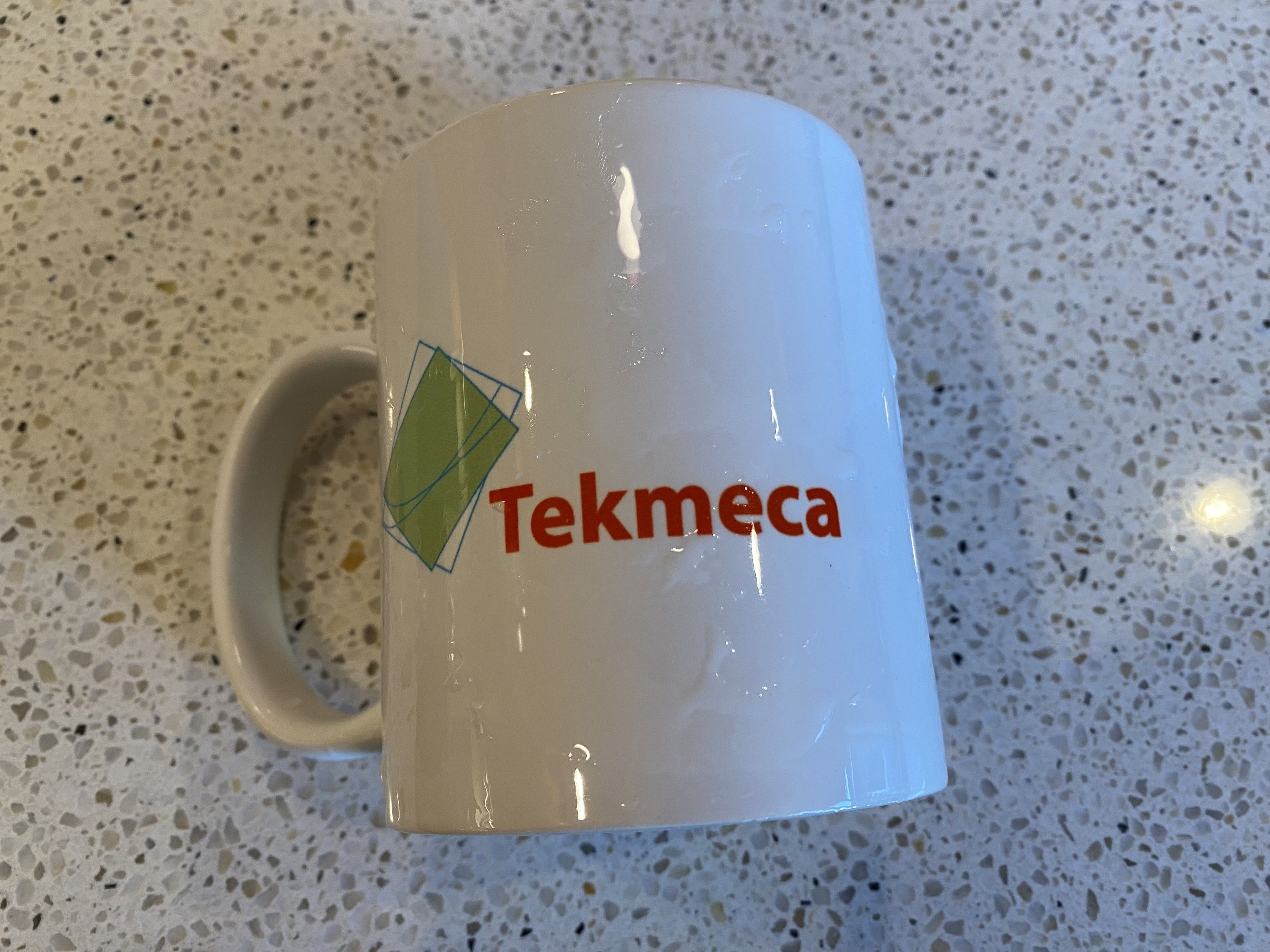 photograph, white corporate coffee mug, company name side “Tekmeca” in red helvetica next to an abstract geometric green and blue logo