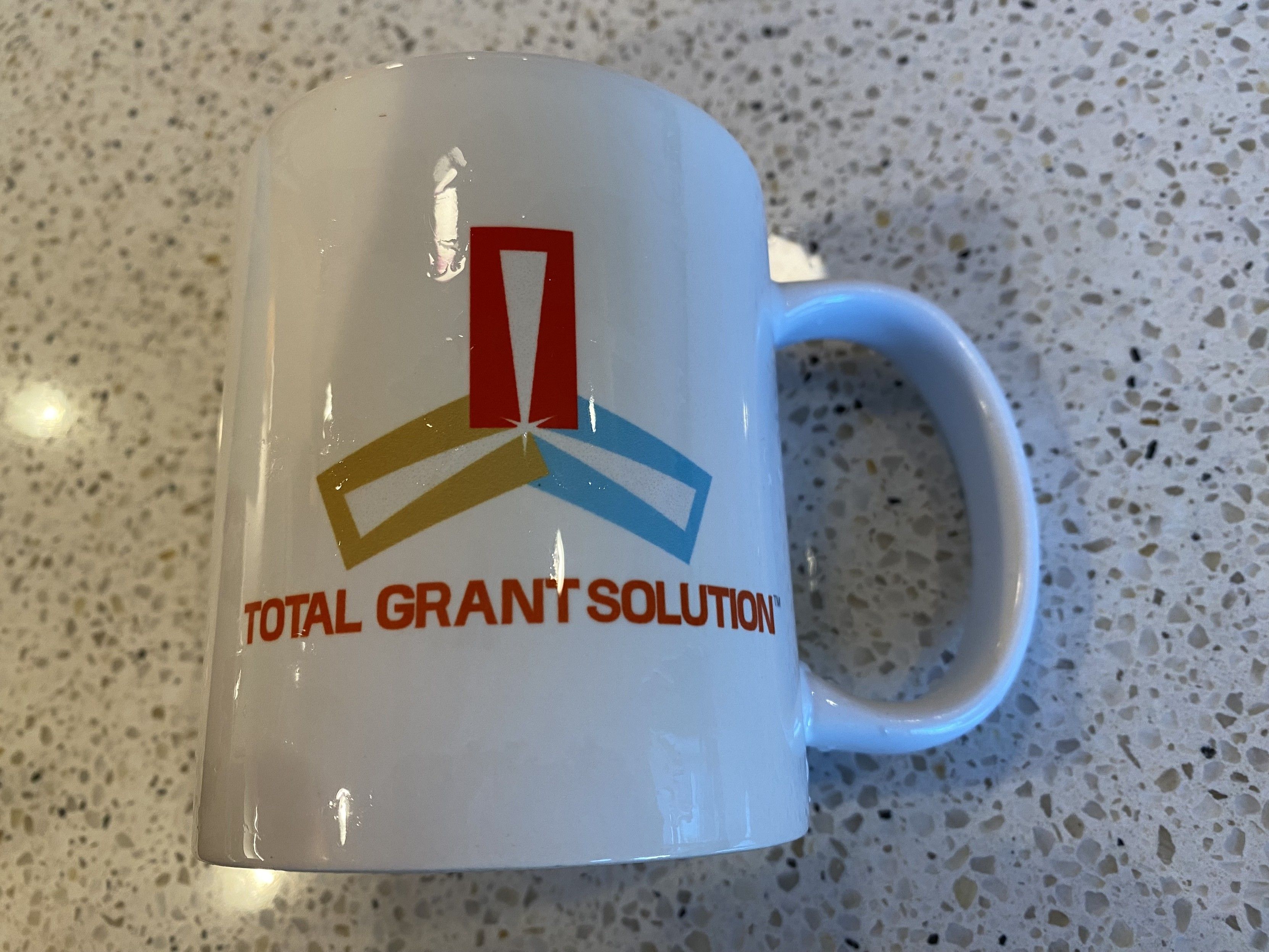 photograph, white corporate coffee mug, product name side “TOTAL GRANT SOLUTION” in red helvetica under to an abstract cut-up looking block and line three point star in red, yellow, and blue