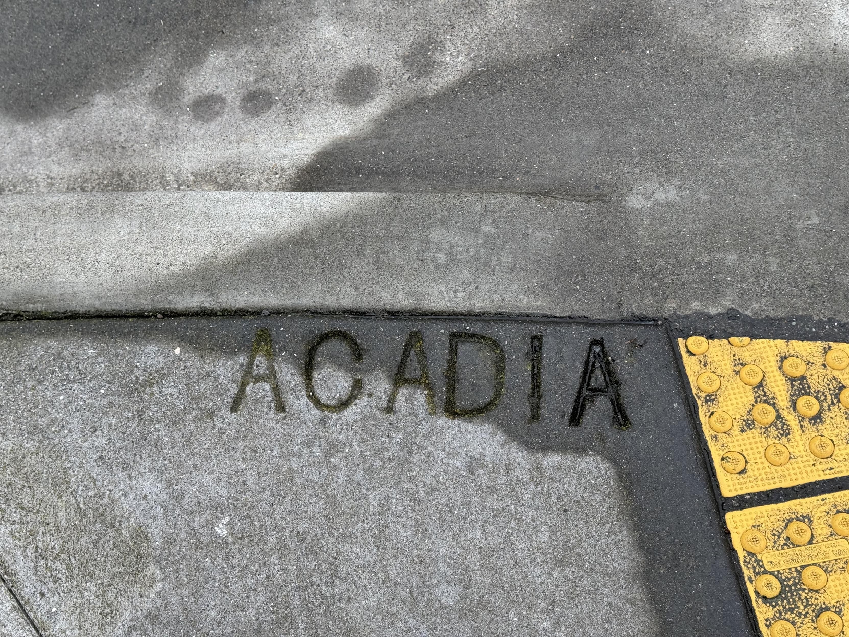a photograph of a concrete sidewalk ending at a concrete curb. to the right are the yellow bumpy ramp sidewalk cut that SF deploys everywhere, and at center, the concrete is stamped with the street name, as SF deploys these stamps at all intersections:ACADIA