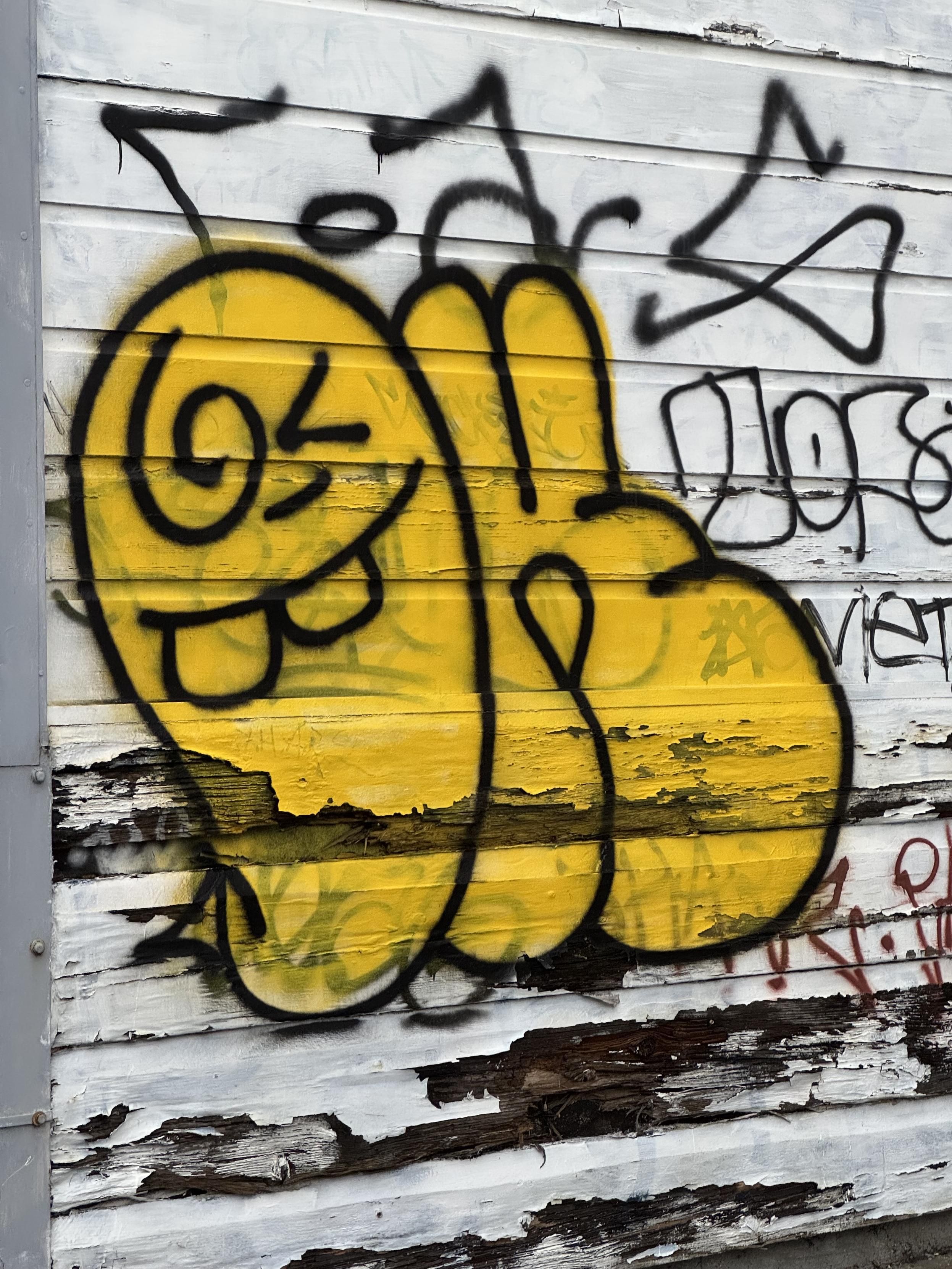 graffiti on the side of a wood-sided garage in Orange Alley, in San Francisco. The garage is tagged up with mostly black nonsense and some red nonsense which has been tagged over with a large, frame filling yellow figure outlined in black, filled in bright yellow. The figure has two buck teeth but mouth closed, a - for an nose, a spiral for one eye and a < for the other eye. Next to the figure is another black lined shape of the same size filled in yellow that could be a ✌️or something else, idk, that’s what I see at a glance.