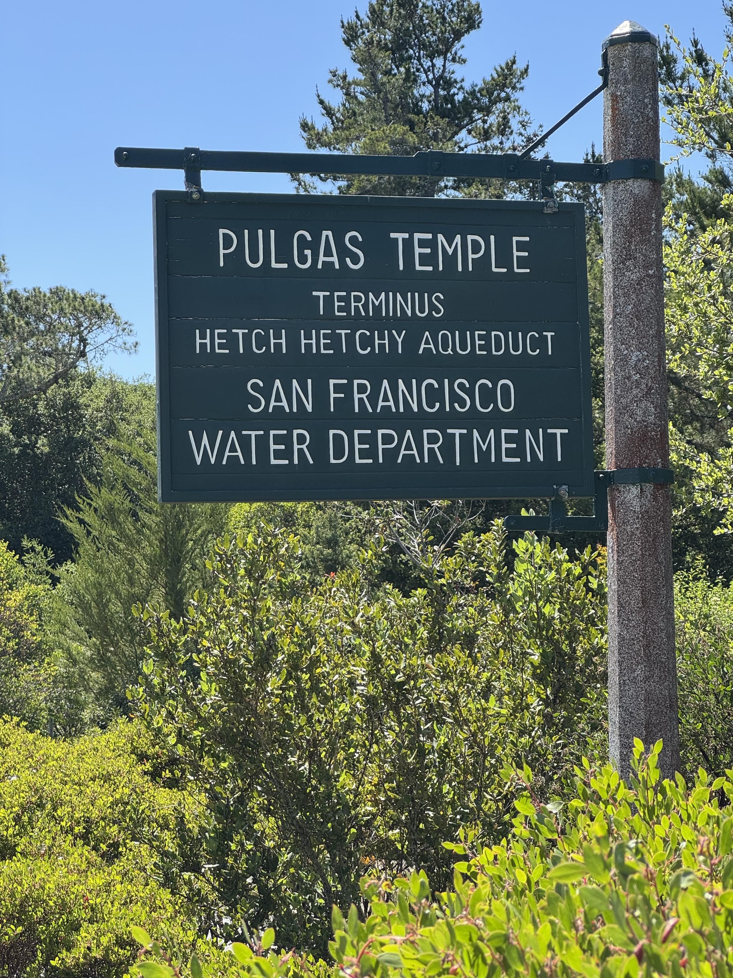 A green sign with white uppercase routed letters hanging from a green pipe on a larger steel pole, surrounded by green deciduous, pine, and bushes under a clear blue sky, which reads:PULGAS TEMPLETERMINUSHETCH HETCHY AQUEDUCTSAN FRANCISCOWATER DEPARTMENT