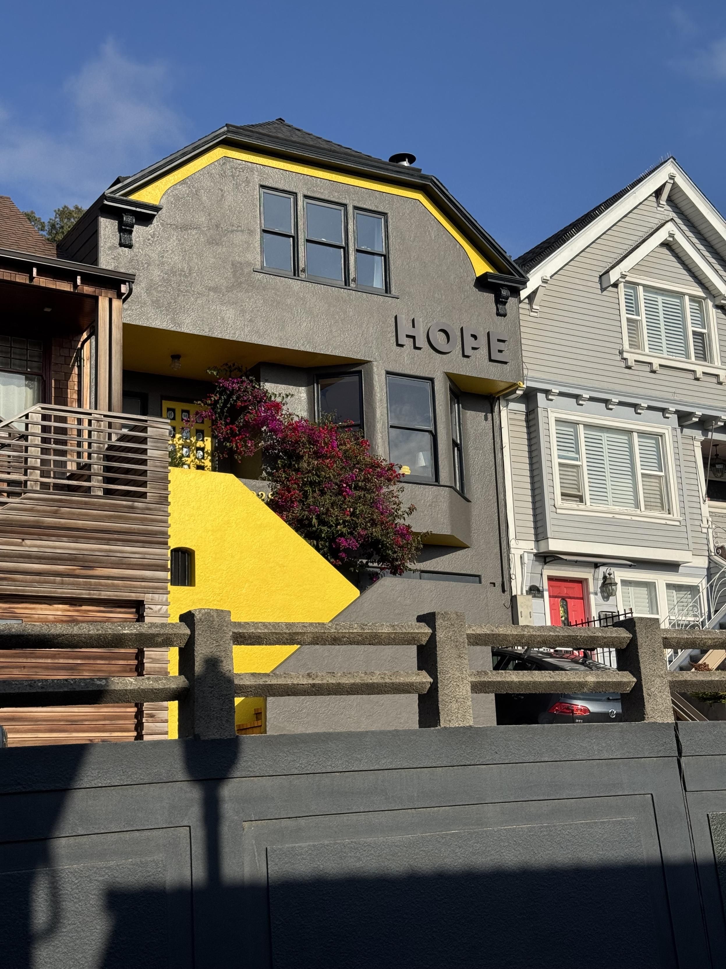 a photograph of the front facade of a house in san francisco. the house is stucco or some other flat surface with a staircase first rising to the right, parallel to the house that then turns and rises to the left parallel to the house. The windows are black aluminum and double-hung with no shutters. The inner wall facade of the lower staircase is bright yellow and presents as an arrow wedge pointing right because the outer facade is the gray again. A decorative yellow line along the barn style roof pitch is also the same yellow.between first first and second story windows, the owner (I presume) has affixed the word HOPE in raised 2’ high all capital san serif letters painted the same dark gray as the house. I don’t know what is going on with that, but I’m getting hippie shit vibes, on the hippie/punk spectrum.Anyway, for more on More Windows (high color), check out https://www.youtube.com/watch?v=xX9B_BYMXQE.
