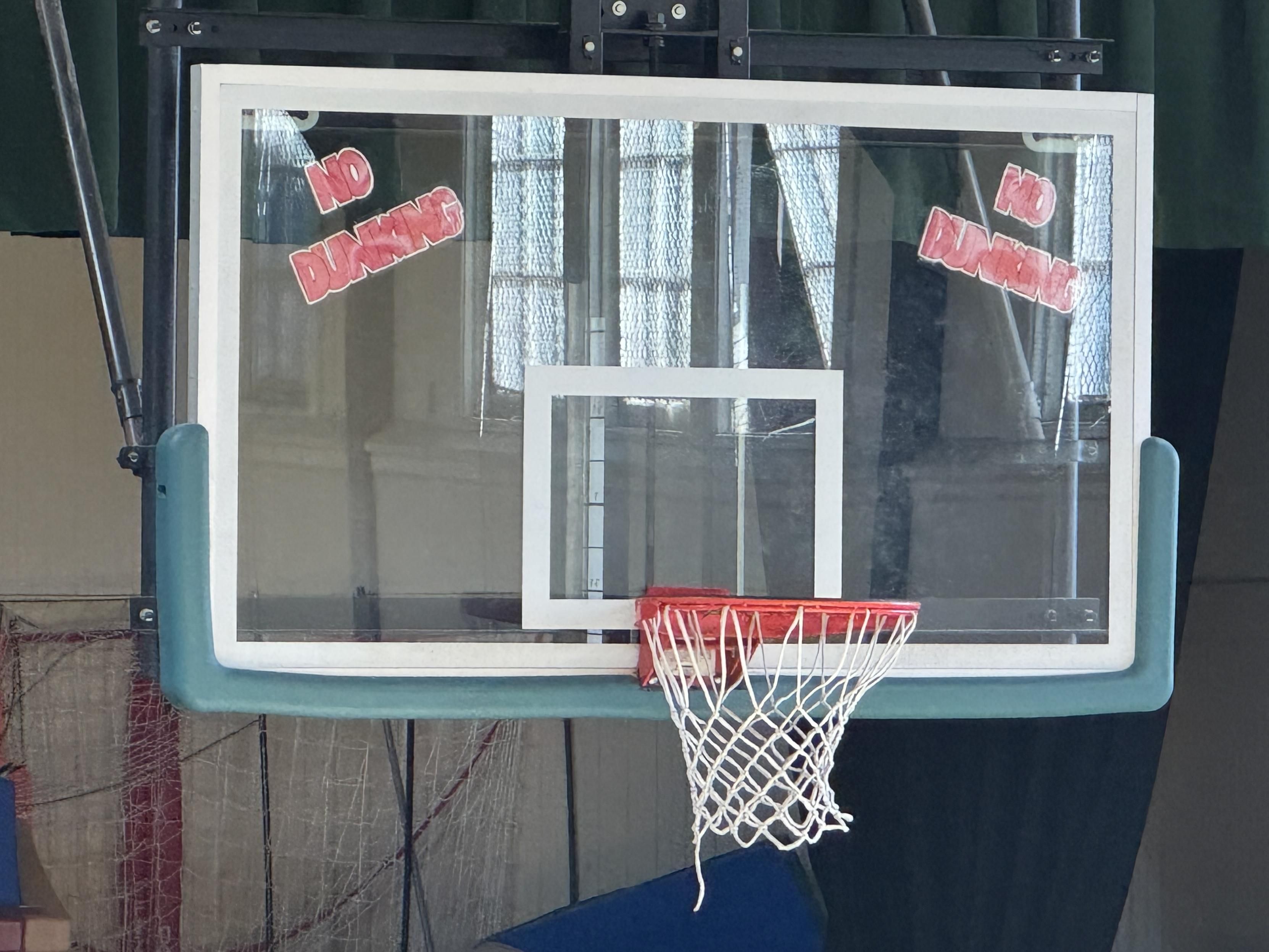 a basketball hoop in the george moscone recreation center in san francisco. the backboard is glass, and the facility has taped hand-made signs that say "NO DUNKING" behind the glass in the top left and right corners.