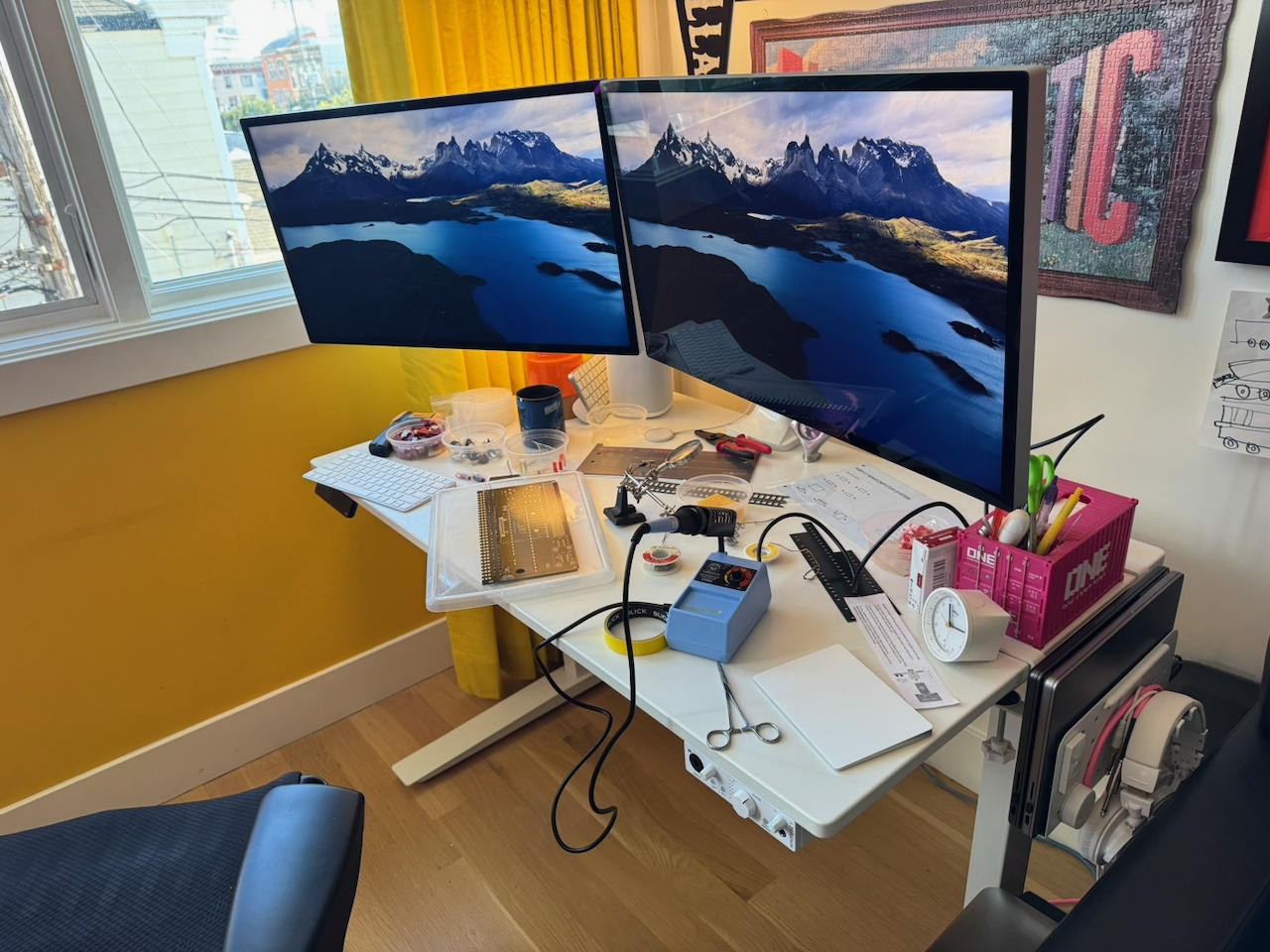 my posting station, two apple studio displays on a white fully standing desk w/ a black embody chair. the desk is covered in entry level soldering gear, coffee cups, an NA beer can, and the kit, in pieces, during assembly.