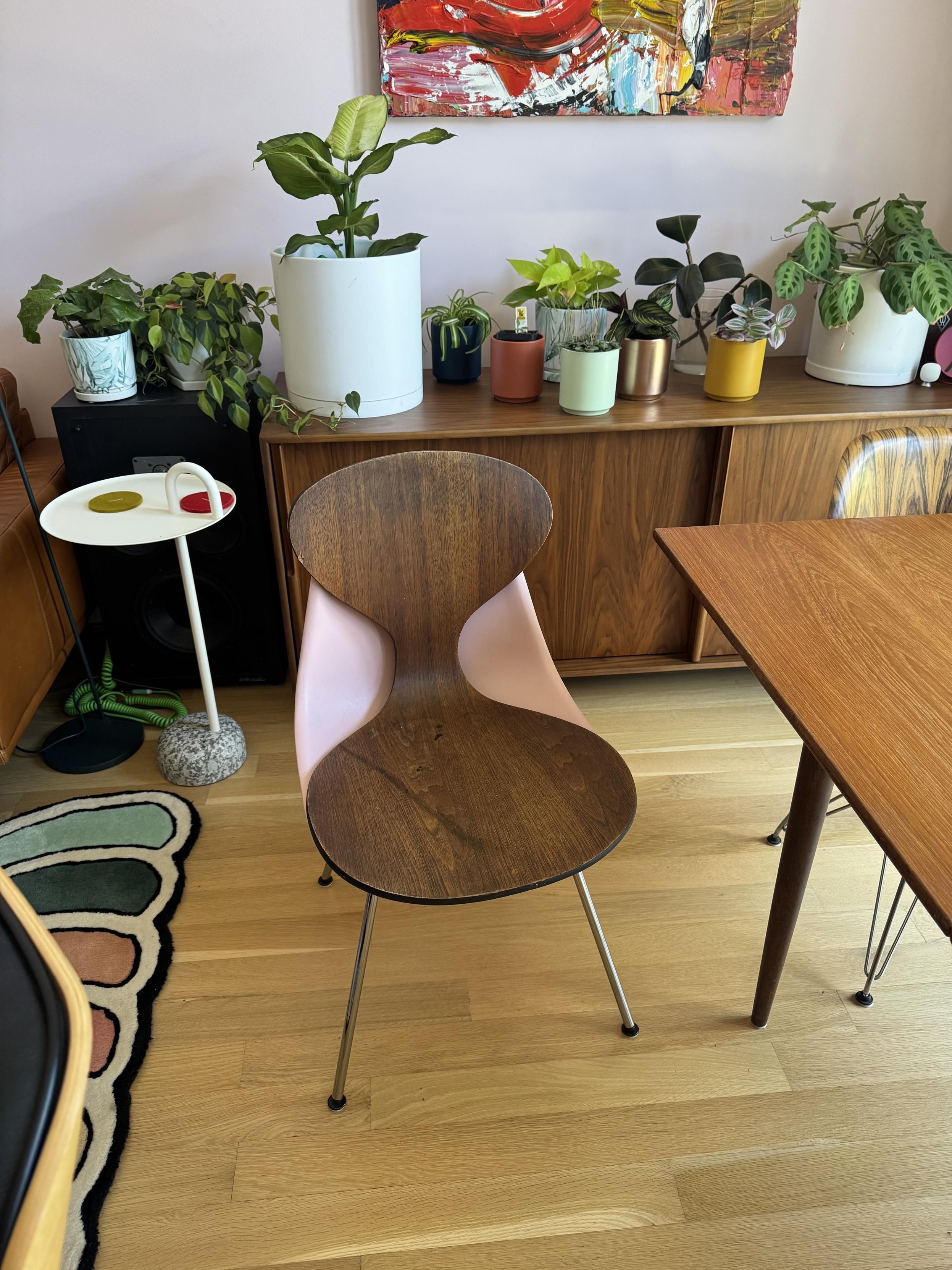 the one-piece bent ply seat and back, resting on a pink eames shell chair on an H-base next to our kitchen table to the right and a Hay side table to the left.A bunch of potted plants are on the credenza on the back wall.
